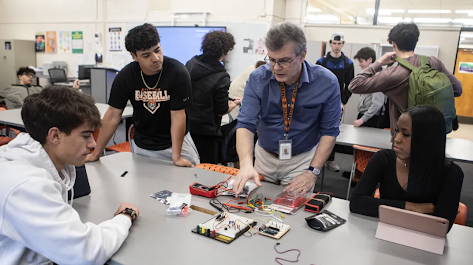 An instructor demonstrates electronic components and a multimeter to a group of high school students gathered around a classroom table.